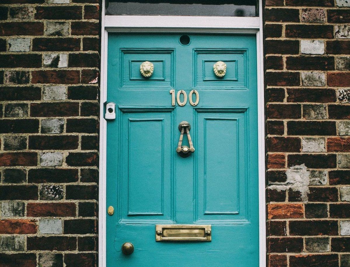 A imagem ilustrativa apresenta uma porta de tonalidade azul claro, com uma parede de tijolos à vista ao redor. A porta contém alguns enfeites dourados, como o número 100, uma fenda para a entrada de correspondência, o trinco redondo, um batedor de porta, uma câmera ao lado e duas cabeças de leões em cima.