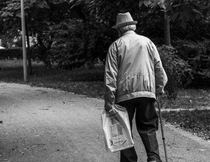 Imagem Ilustrativa. Está em preto e branco, de um senhor de costas, de jaqueta e chapéu, caminhando em uma calçada de um parque, com uma bengala em uma mão, e um jornal na outra.