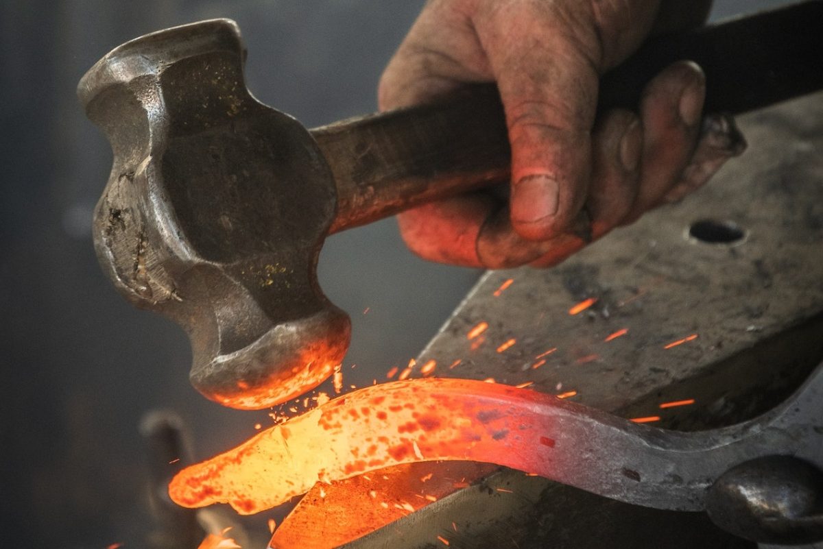 A imagem mostra uma ferradura incandescente sendo forjada com um martelo em uma bigorna.