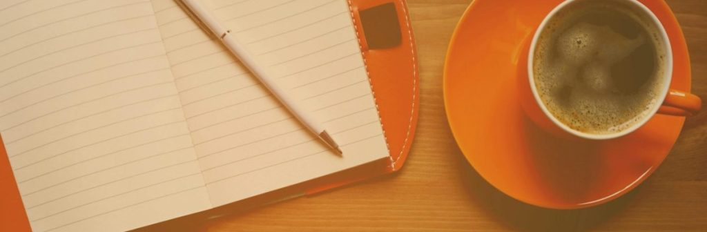 Foto de uma xícara de café, e um caderno com um lápis, sobre uma mesa de madeira.