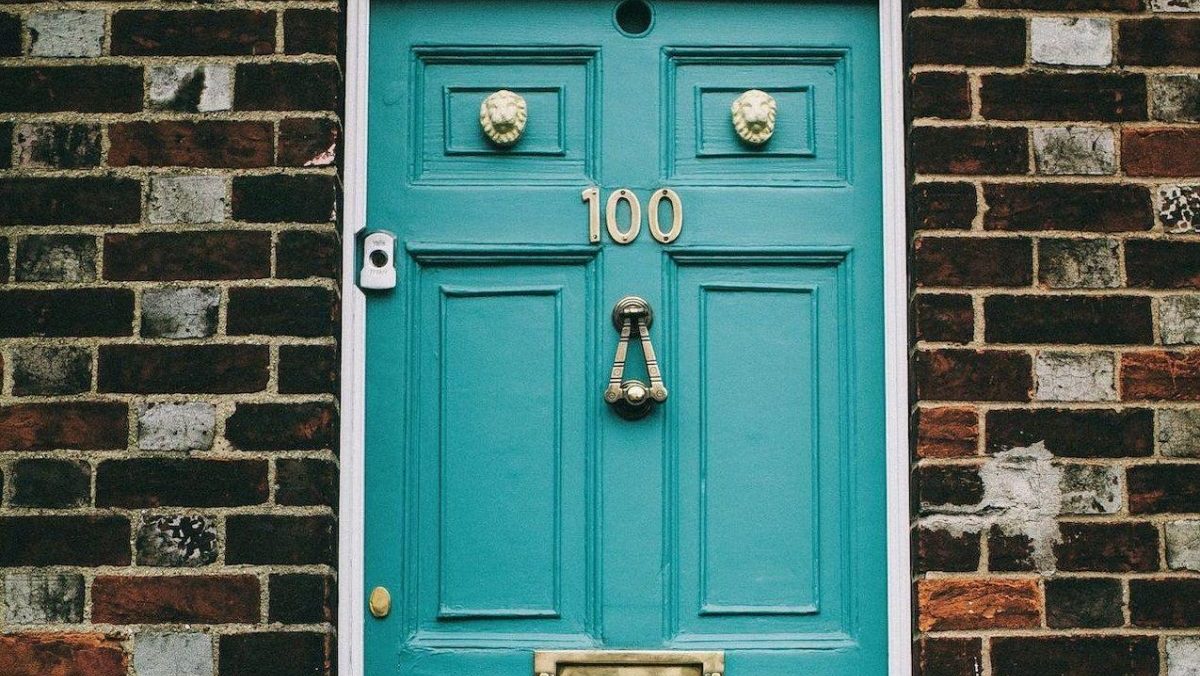 A imagem ilustrativa apresenta uma porta de tonalidade azul claro, com uma parede de tijolos à vista ao redor. A porta contém alguns enfeites dourados, como o número 100, uma fenda para a entrada de correspondência, o trinco redondo, um batedor de porta, uma câmera ao lado e duas cabeças de leões em cima.